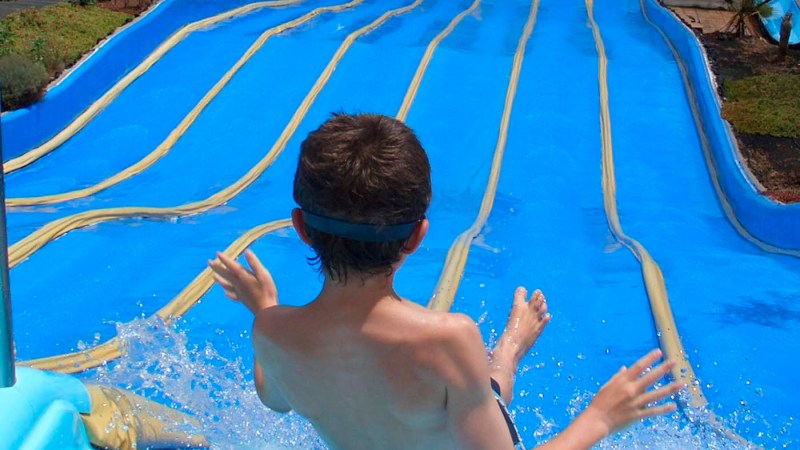 Child sliding down a blue water slide into a pool with people swimming.