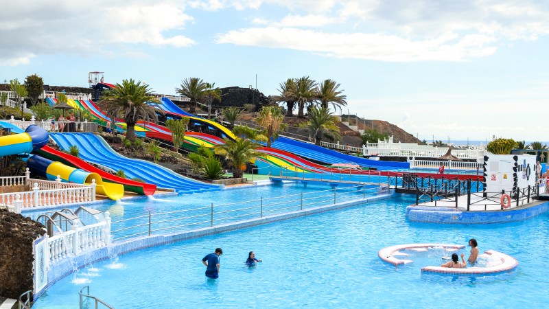 Water park with colorful slides and people swimming in a large pool under a partly cloudy sky.