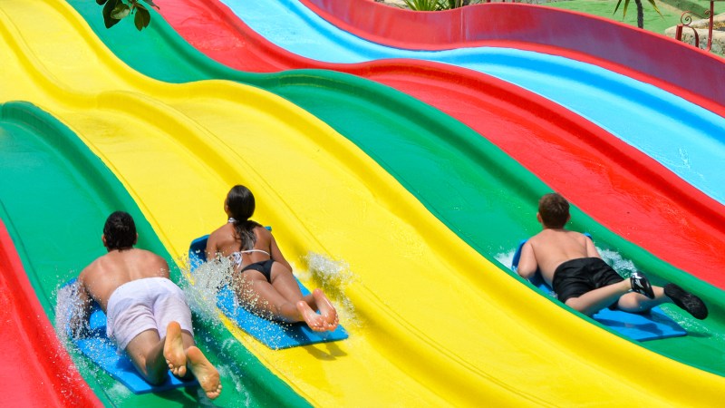 Three people on mats sliding down colorful water slides with a pool in the background.