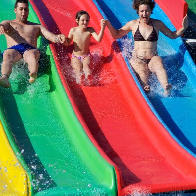Three people on colorful water slides holding hands, splashing into a pool.