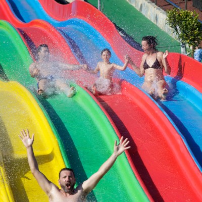 Three people sliding down colorful water slides, splashing water and smiling in excitement.