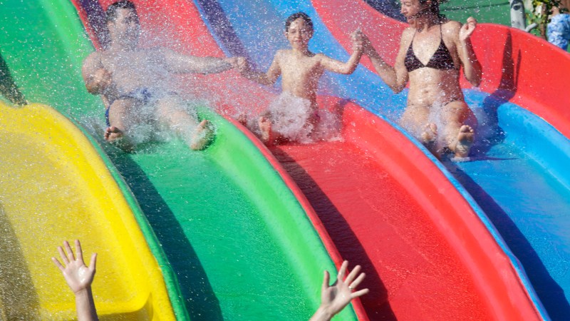 Three people sliding down colorful water slides, splashing water and smiling in excitement.