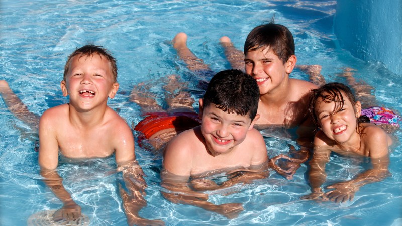 Four children smiling and playing in a swimming pool on a sunny day.