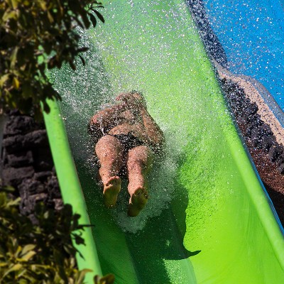 Person sliding down a green waterslide, splashing water, with plants on the side.