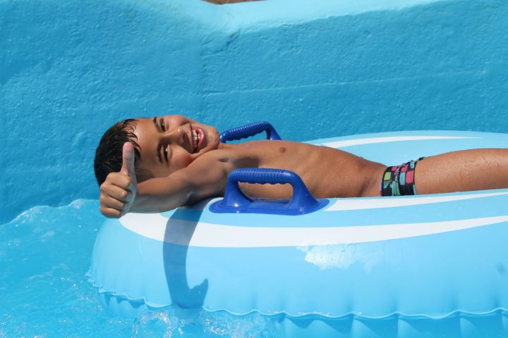 Smiling child on blue inflatable ring gives thumbs up in pool.